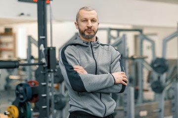 Portrait of sporty man with folded hands in the gym, handsome bearded trainer looking at the camera