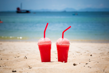 Two watermelon smoothies in disposable plastic cups with tubes. On a sandy tropical beach. Against the backdrop of the blue sea. Healthy lifestyle and refreshing