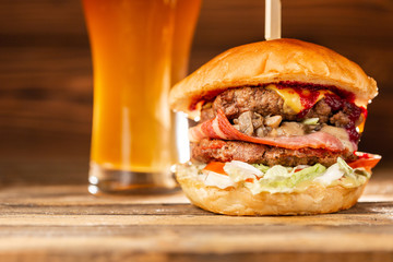 A big burger with two beef cutlets, cooked on charcoal. Lunch with Glass of beer on a wooden background. The concept of fast food and unhealthy food