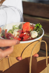 concept takeaway dishes, fresh salad greens, tomatoes and sprouted grains in paper plate in the hands of a young girl who eats it outdoors
