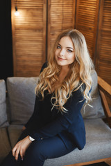 Fototapeta premium Beautiful and fashionable young blond business lady with blue eyes in a stylish dark blue suit sits on a sofa and posing at loft interior