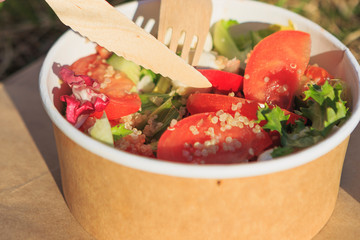 Woman eating healthy salad from paper container over green grass with flying hair on a sunny spring day. backlight