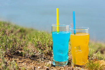two glasses with colored drinks and with straws stand in nature, against the background of blue water, the concept of traveling together