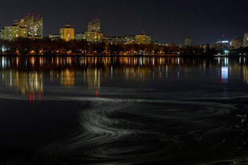 Obraz premium dark cityscape with illuminated buildings, waves on river and night sky