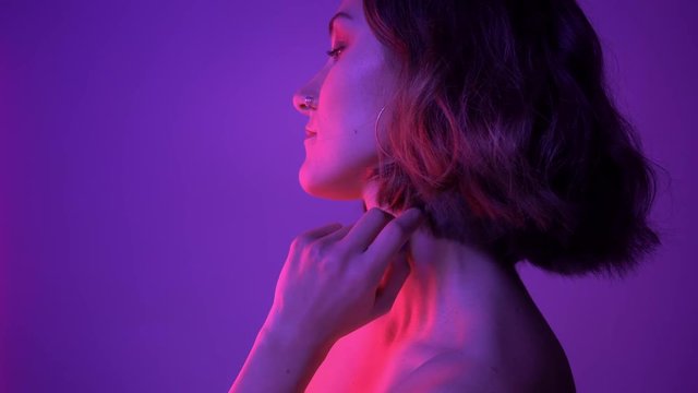 Closeup side view shoot of young alluring female posing and fixing her hair in front of the camera with neon purple background