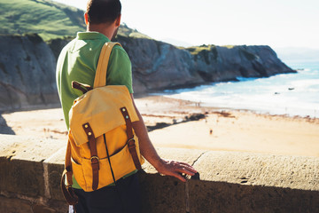 tourist traveler with yellow backpack standing on green top on mountain, hiker view from back looking on ocean, man enjoying nature panoramic landscape in trip, relax holiday concept