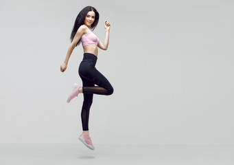 Fototapeta na wymiar Amazing woman in trendy sportswear jumping. Smiling beautiful slim brunette young girl in fashion leggings and pink top expressing happy emotions. Gray background. Sport, fitness, lifestyle concept.