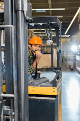 good-looking indian warehouse worker sitting in forklift machine and talking on walkie talkie