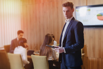Stylish young businessman wearing a jacket and a shirt on the background of a working office with people working with a tablet
