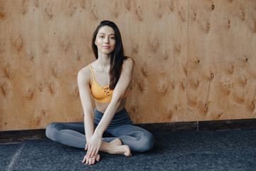 Young attractive fitness girl sitting on the floor near the window on the background of a wooden wall, resting on yoga classes