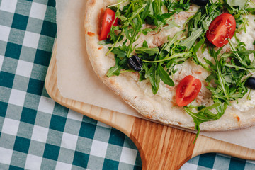 Neapolitan pizza with tuna, cheese, arugula, basil, tomatoes, olives, sprinkled with cheese on a wooden table on a tablecloth in a cell with a place for the text.