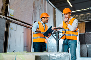 upset warehouse worker with pallet jack standing near angry yelling indian colleague