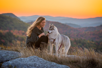 Woman smiles looking her husky dog sitting on an open ridge with the sun rising behind her and mountain ridges. - obrazy, fototapety, plakaty
