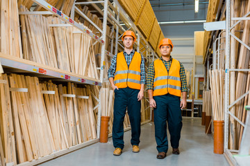 Fototapeta premium multicultural warehouse workers in safety vasts and helmets walking along racks with wooden construction materials