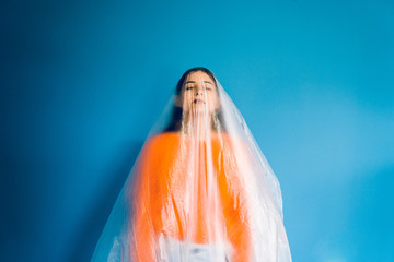 Portrait of young woman in orange in front of blue wall covered with nylon plastic bag, antipollution pollution save the planet oceans ecology concept