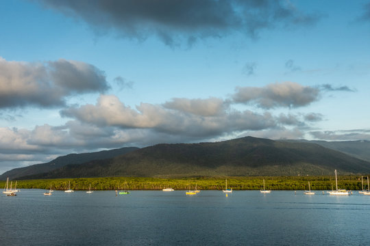 Cairns, Australia - February 18, 2019: Evening shot at sunset. Anchored white small sailing boats on Chinaman Creek under heavy cloudscape. Backdrop is rainforest hills.
