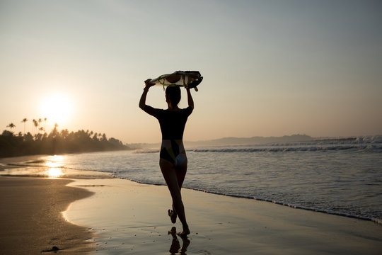 Surfer girl walking with surfboard on the beach at sunrise