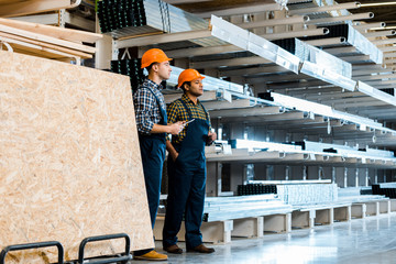 Fototapeta premium two multicultural friends in uniform standing near racks with metallic construction materials