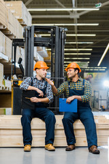 two smiling multicultural workers with clipboards talking while sitting on plywood