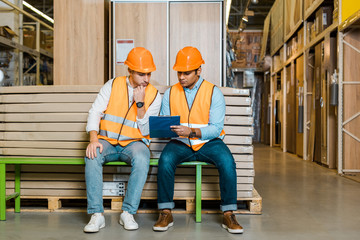 concentrated multicultural workers sitting on bench in warehouse and looking at clipboard