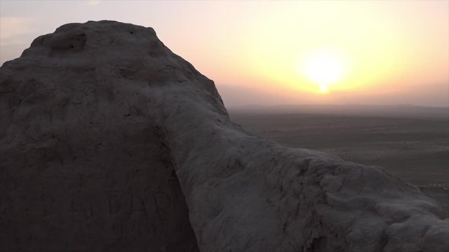 A medium, still shot of the ruins of an old wall made of clay with feint writing on it and the sunset in the background