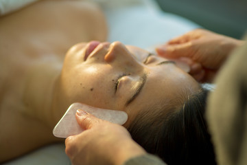 Young woman receives facial rejuvenation with gua sha rose quartz in spa wellness center