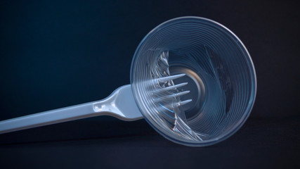 Garbage from disposable tableware. A plastic fork pierces a plastic cup on a black background. Eco concept and injunction on the use of plastic flatware. Copy space.