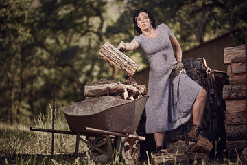 A pergnant woman in a dress throwing wood into a wheelbarrow on a ranch in Northern California.