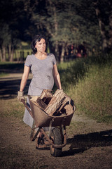 A pregnant woman in a vintage dress and work boots pushing a wheelbarrow full of chopped wood on a rural ranch in Northern California.