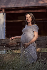 A dirt covered, pregnant woman in a dress  resting against a fence on a ranch in rural Northern California