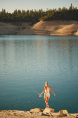 A young, barefoot woman in a swimwear coverup walking down to a lake while holding a fiishing pool on a summer day.