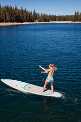 A young woman in a blue bikini doing a pivot turn on a paddle board while paddling across a small lake in the mountains of Northern California.