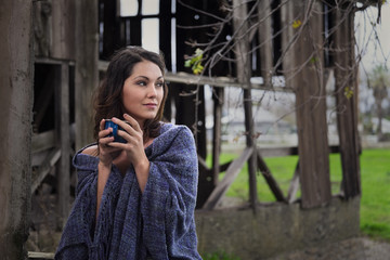 Fototapeta premium A farm girl wrapped in a blue shawl drinking a cup of coffee next to an old barn on a small rural farm.
