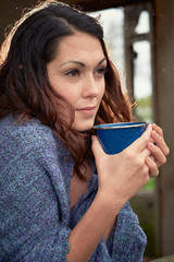 A young woman wrapped in a shawl drinking coffee in an abandoned building of a small, rural farm.