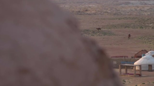 A wide panning shot from behind a rock to a landscape in Uzbekistan with a row of different colored huts, a dry landscape and some camels