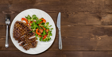 Grilled steak with vegetables on a plate with fork and knife on wooden background. Empty space for text