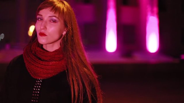 The girl walks in the evening city. Neon lighting. Wearing a black polto and a burgundy scarf.