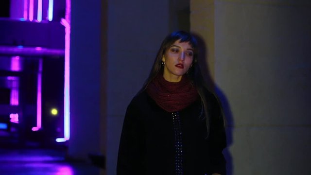 The girl walks in the evening city. Neon lighting. Wearing a black polto and a burgundy scarf.