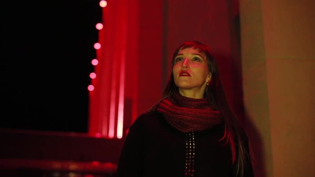The girl walks in the evening city. Neon lighting. Wearing a black polto and a burgundy scarf.