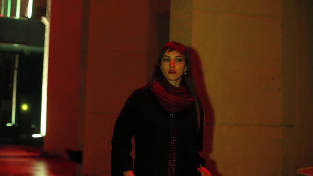 The girl walks in the evening city. Neon lighting. Wearing a black polto and a burgundy scarf.