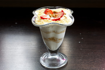 Dessert in a glass goblet. Layers laid biscuit crumbs and cream. Decorated with strawberry slices. With the addition of walnut. On a dark background.