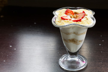 Dessert in a glass goblet. Layers laid biscuit crumbs and cream. Decorated with strawberry slices. With the addition of walnut. On a dark background.