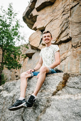A man sitting on background of the large  rocks wall on a big stone. full length, view from down side. looking at the camera. Close up. Place for text and design. Man poses at cliff near mountain.