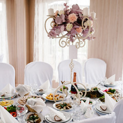 A composition of flowers and green is on a festive table at the wedding banquet hall. Stands candles. Close up.