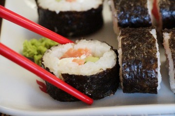 Red shopsticks holds maki sushi (makizushi, norimaki, makimono or sushi rolls) with salmon and cucumber on white plate.  Sushi, Japanese cuisine, Food and drink concept.