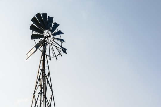 Low angle view of an old-fashioned, multi-bladed, metal wind pump atop a lattice tower in backlight against a pale blue sky.