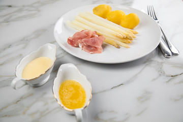 Weißer Spargel mit Sauce Hollandaise, zerlassener Butter, geräucherter Schinken und Kartoffeln auf Teller und Marmor Hintergrund