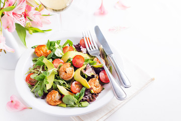 Delicious fresh salad with prawns, greens, cucumber, avocado and tomato. with white wine and flowers on light background
