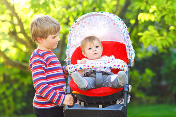 Little blond kid boy playing with baby sister. Happy siblings in garden. Baby girl sitting in pram...