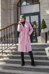 Stylish Muslim girl with a scarf on her head in a pink coat in the city. Stylish headband. The image of a modern woman in the city on the background of architecture